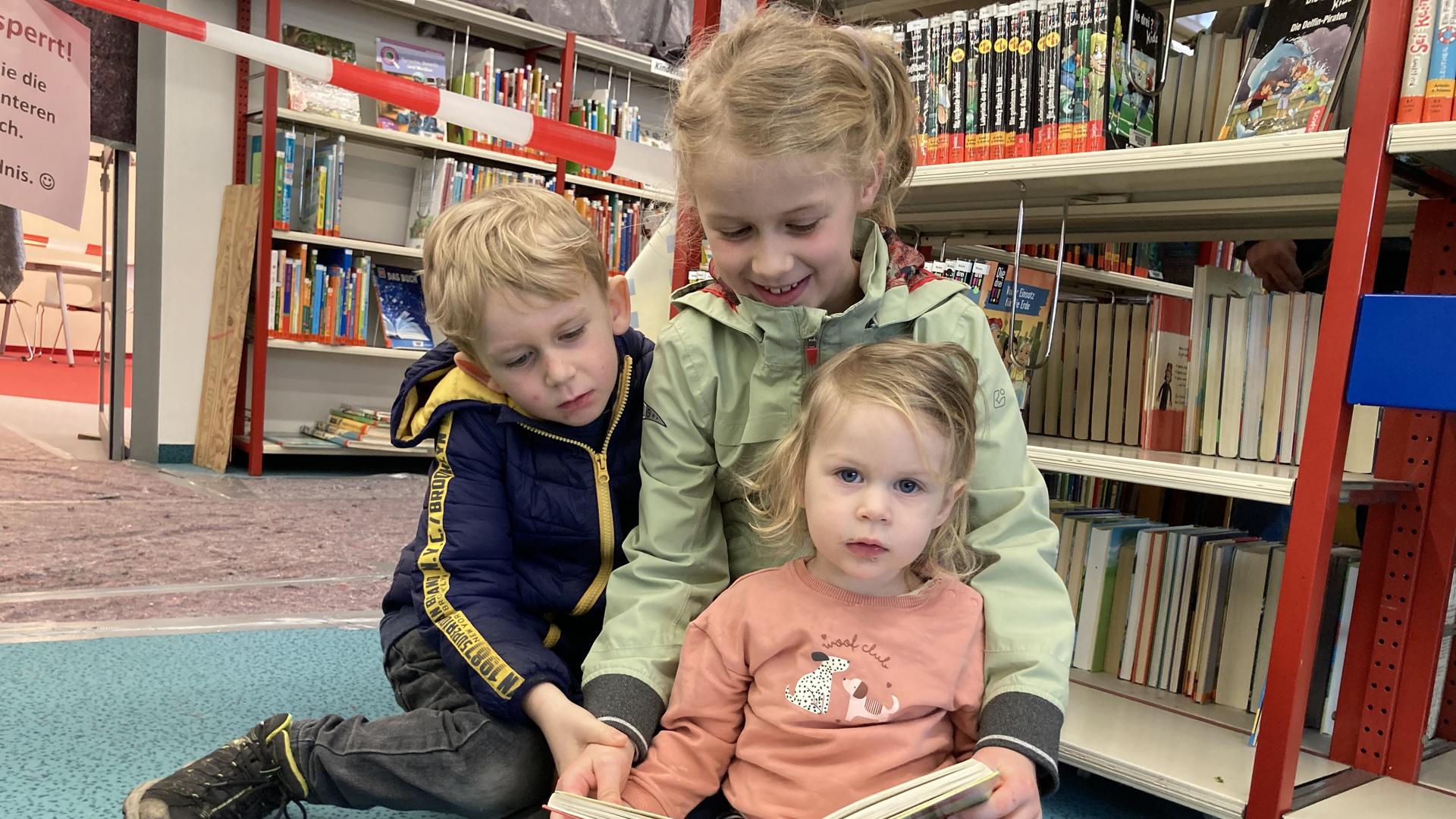 drei Kinder sitzen in einer Bibliothek und lesen ein Buch