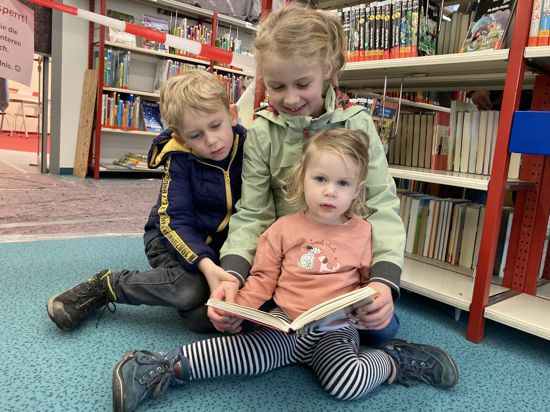drei Kinder sitzen in einer Bibliothek und lesen ein Buch