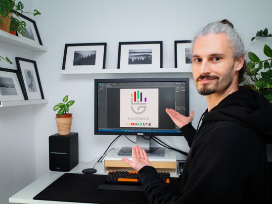 ein Mann sitzt an einem Schreibtisch mit Computer, auf dem Monitor ist ein Logo zu sehen