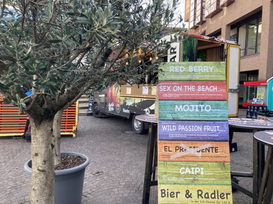 Olivenbäume im Vordergrund, dazu ein buntes Schild mit Cocktailnamen, im Hintergrund ein Imbisswagen