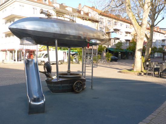Ein Spielgerät in Form eines metallenen Zeppelins mit einer Rutsche dran und einem Ausguck steht auf einem Platz in einer Stadt