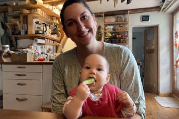 ein Baby sitzt auf dem Schoß seiner Mutter, es hält ein Stück Paprika in der einen Hand und schiebt sich mit der anderen einen Gurkenstick in den Mund