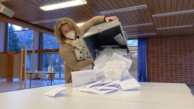 Eine Frau mit Mundschutz kippt den Inhalt einer Wahlurne auf einen Tisch