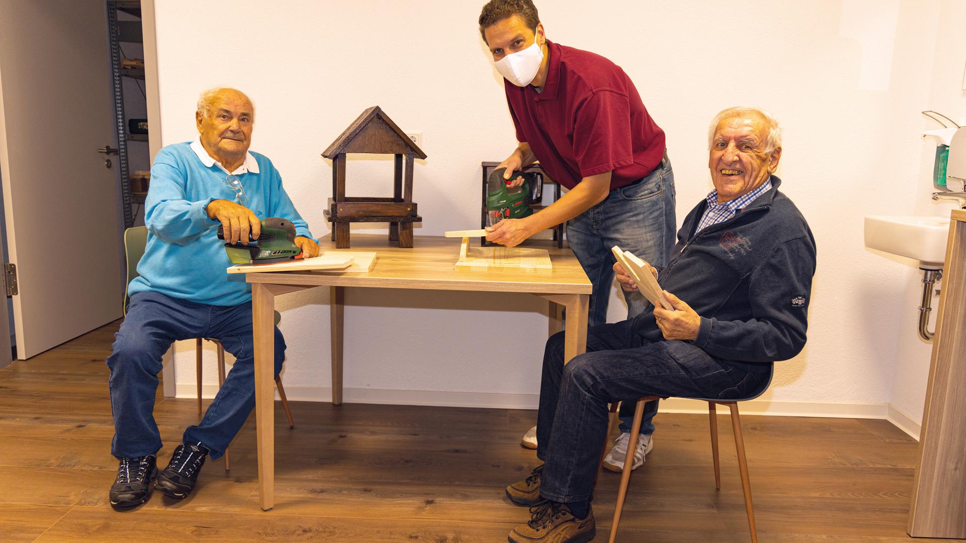 Sid Finner (Mitte) und zwei Senioren bearbeiten an einem Tisch Holz mit Geräten zum Bau von Vogelhäuschen