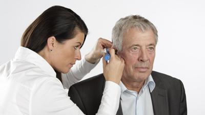 Eine Frau untersucht mit einem Spezialgerät das rechte Ohr eines älteren Mannes.