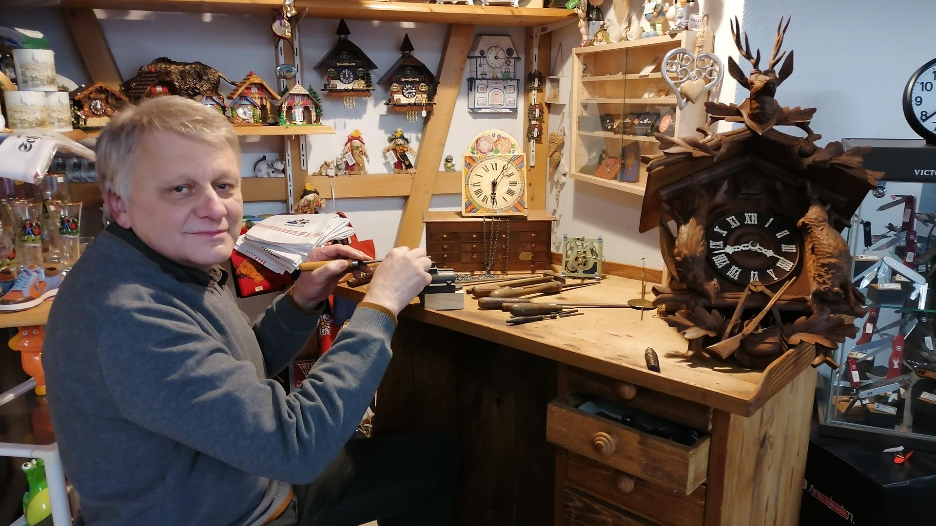 Markus Schleicher sitzt an der Werkbank seines Urgroßvaters. Auf der Werkbank liegen eine traditionelle Kuckucksuhr aus Holz und eine Schwarzwälder Schilduhr.
