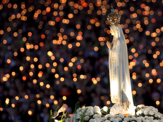 Eine Marienstatue, weiß mit goldener Krone und zum Gebet gefalteten Händen, wird bei einer Prozession im Dunkeln durch eine Menschenmenge getragen, die Lichter in die Höhe hält