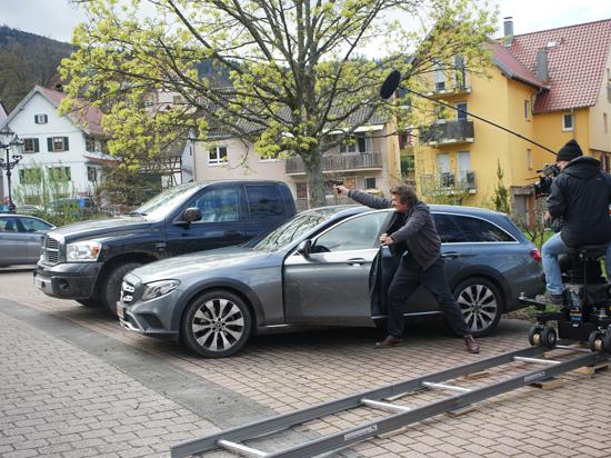 Ein Schauspieler mit einer Pistole steht hinter einem Auto. 