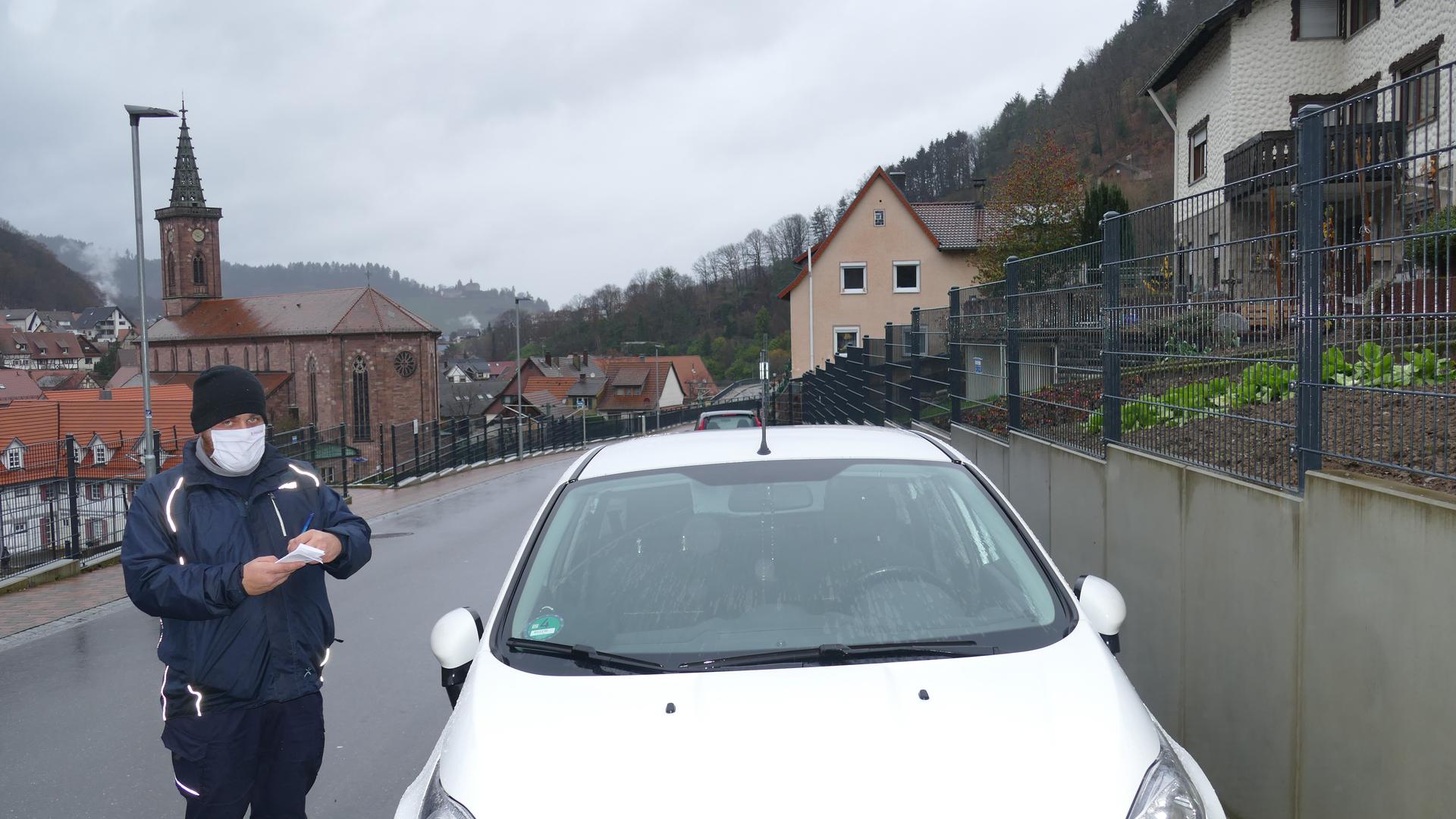 Alexander Preis, Mitarbeiter des Gemeindevollzugsdienst, steht in Weisenbach auf der Straße neben einem weißen Auto, das entgegen der Fahrtrichtung parkt. 