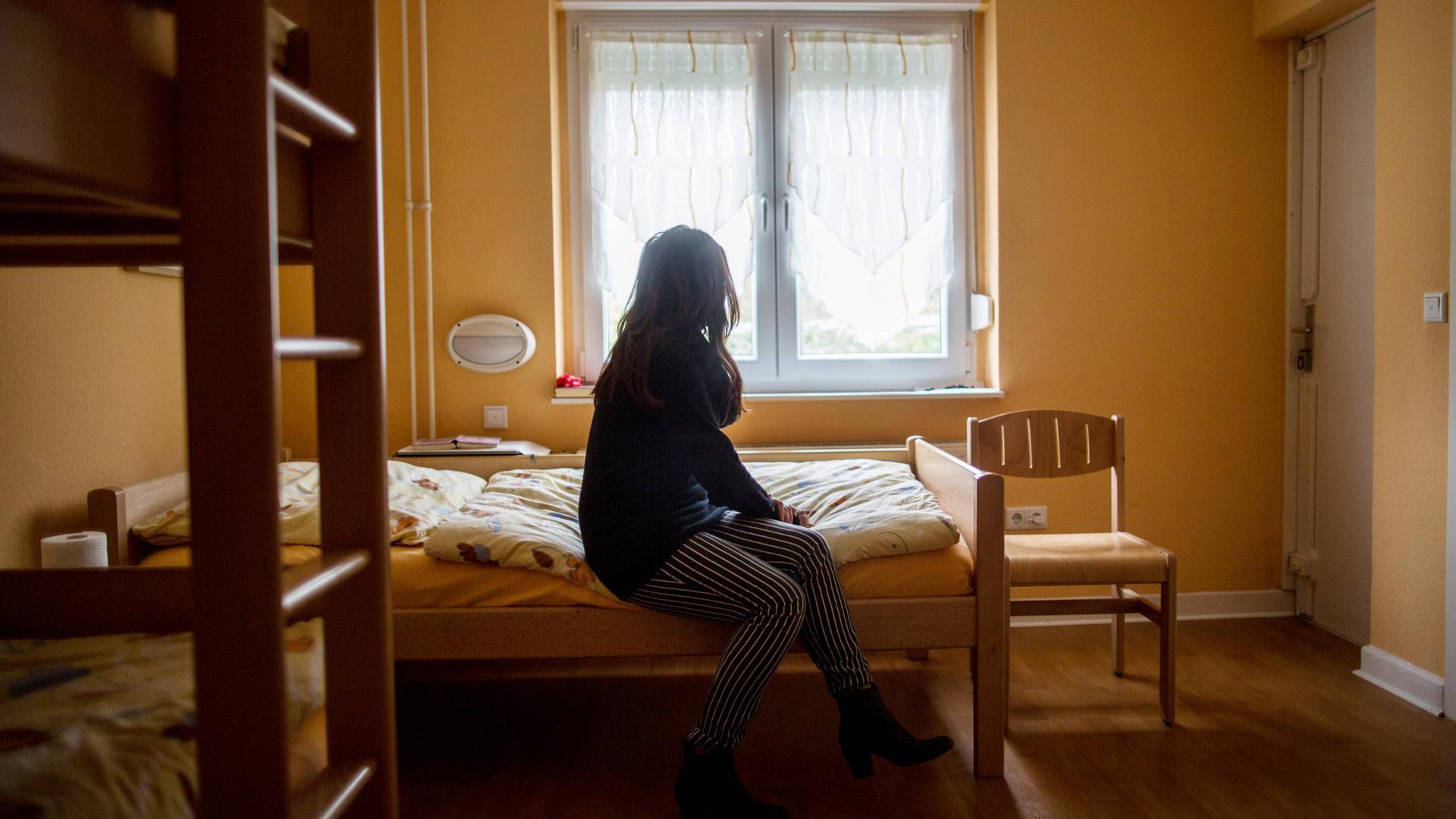 Eine Frau sitzt auf einem Bett vor einem geschlossenen Fenster. Links ist eine Leiter zu einem Etagenbett zu sehen.