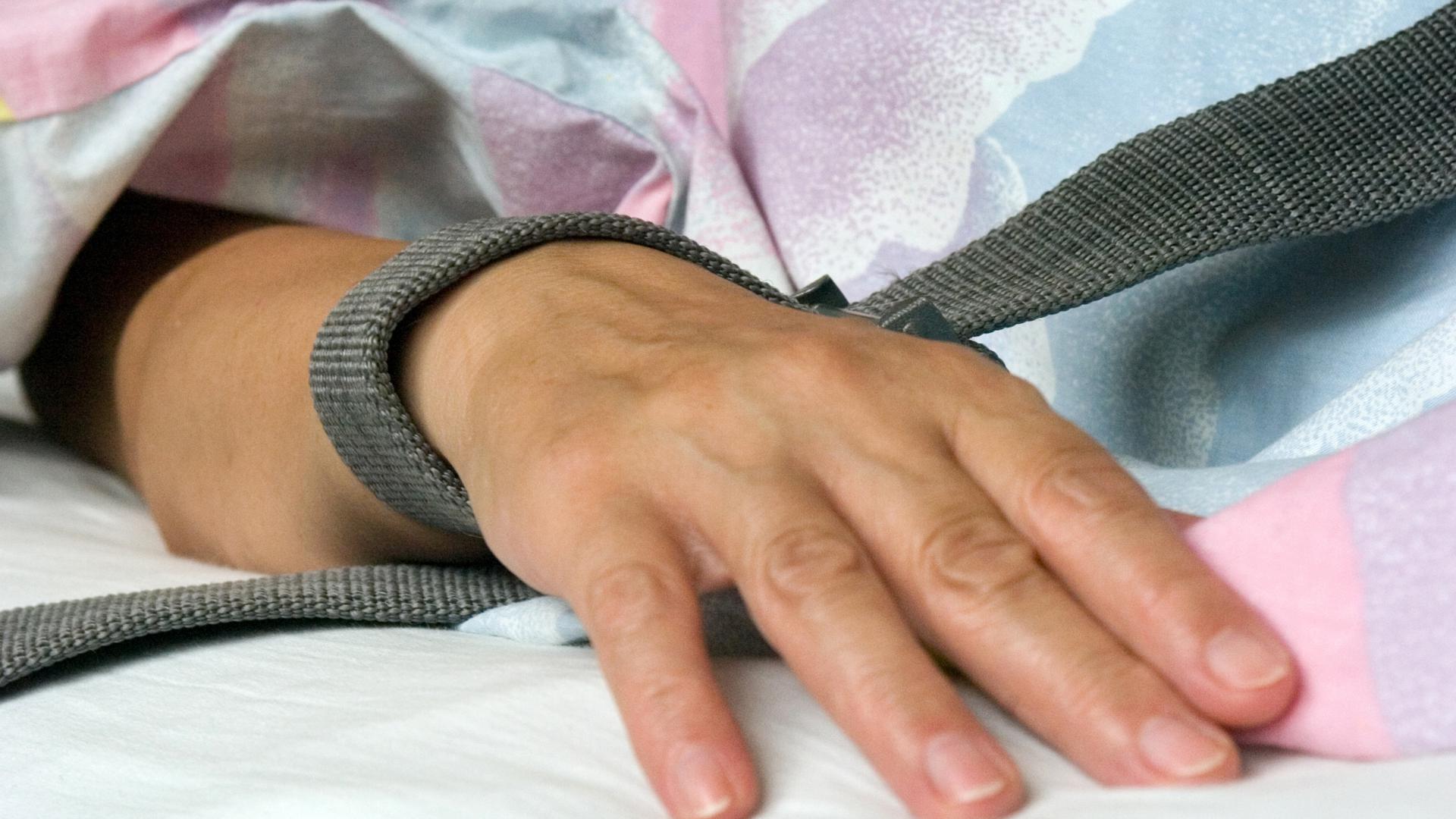 Eine mit einem Textilband festgebundene Hand eines Patienten - die Fixierung bzw. Fixation eines Patienten in der Krankenpflege durch Festschnallen am Handgelenk. 