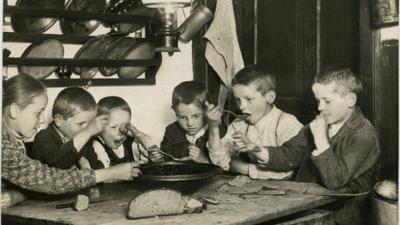 Kinder, die in der Küche an einem Holztisch sitzen und gemeinsam aus einer Schüssel essen, sind auf einer Postkarte des Weltpostvereins zu sehen. Auf dem Tisch liegt ein halber Brotlaib. 