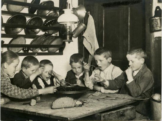 Kinder, die in der Küche an einem Holztisch sitzen und gemeinsam aus einer Schüssel essen, sind auf einer Postkarte des Weltpostvereins zu sehen. Auf dem Tisch liegt ein halber Brotlaib. 