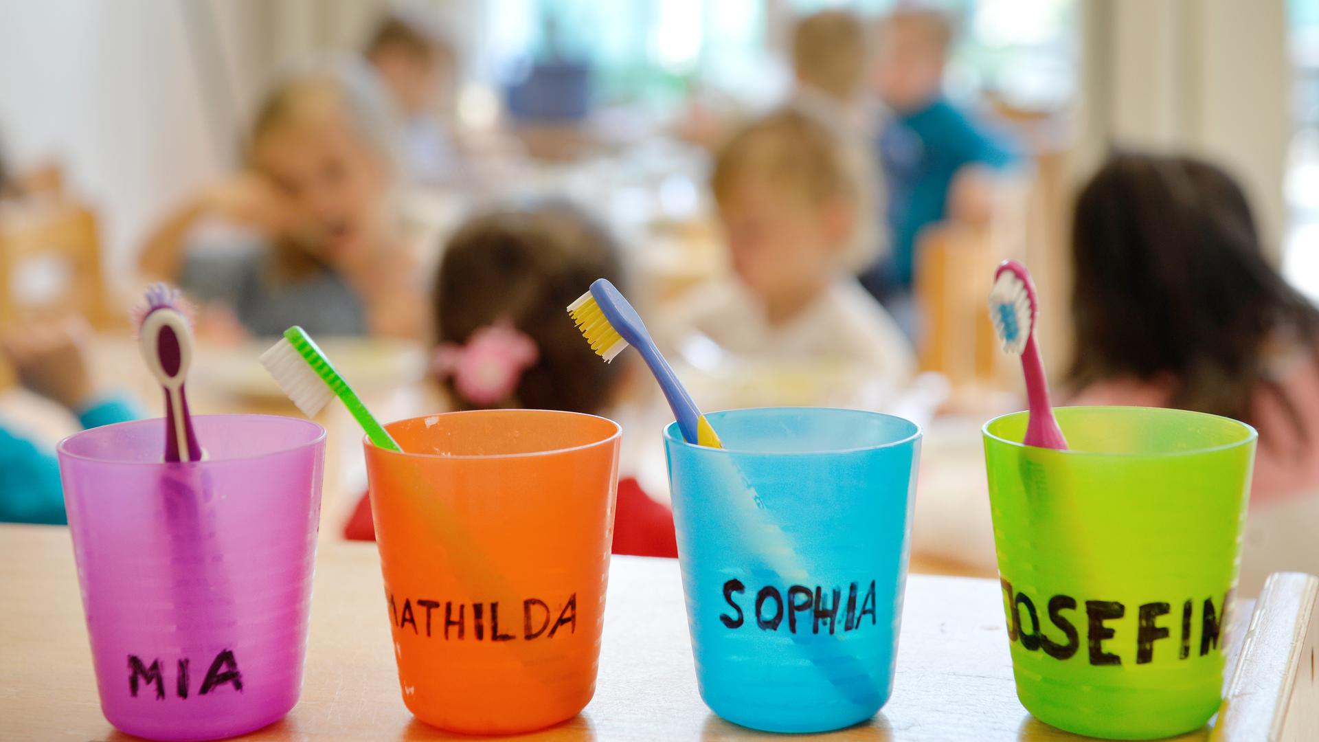 Zahnbürsten und Zahnputzbecher mit den Vornamen der Kinder stehen in einer Kindertagesstätte auf einem Tisch. 