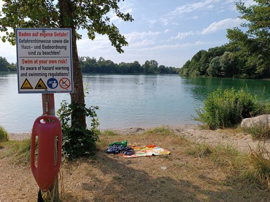 Ein See, am Ufer liegt ein Handtuch und steht ein Schild mit der Aufschrift: „Baden auf eigene Gefahr!“