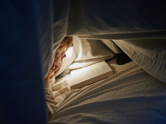 Ein Kind liegt auf einem Bett in einem dunklen Zimmer und liest unter einer Bettdecke mit einer Lampe ein Buch. 