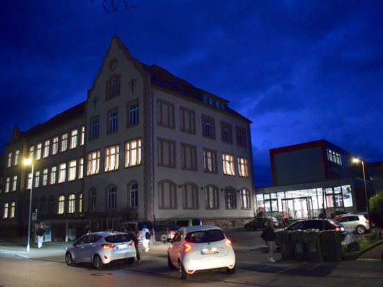 Ein Schulgebäude mit erleuchteten Fenstern in der Dunkelheit, davor Schüler und rangierende Autos.