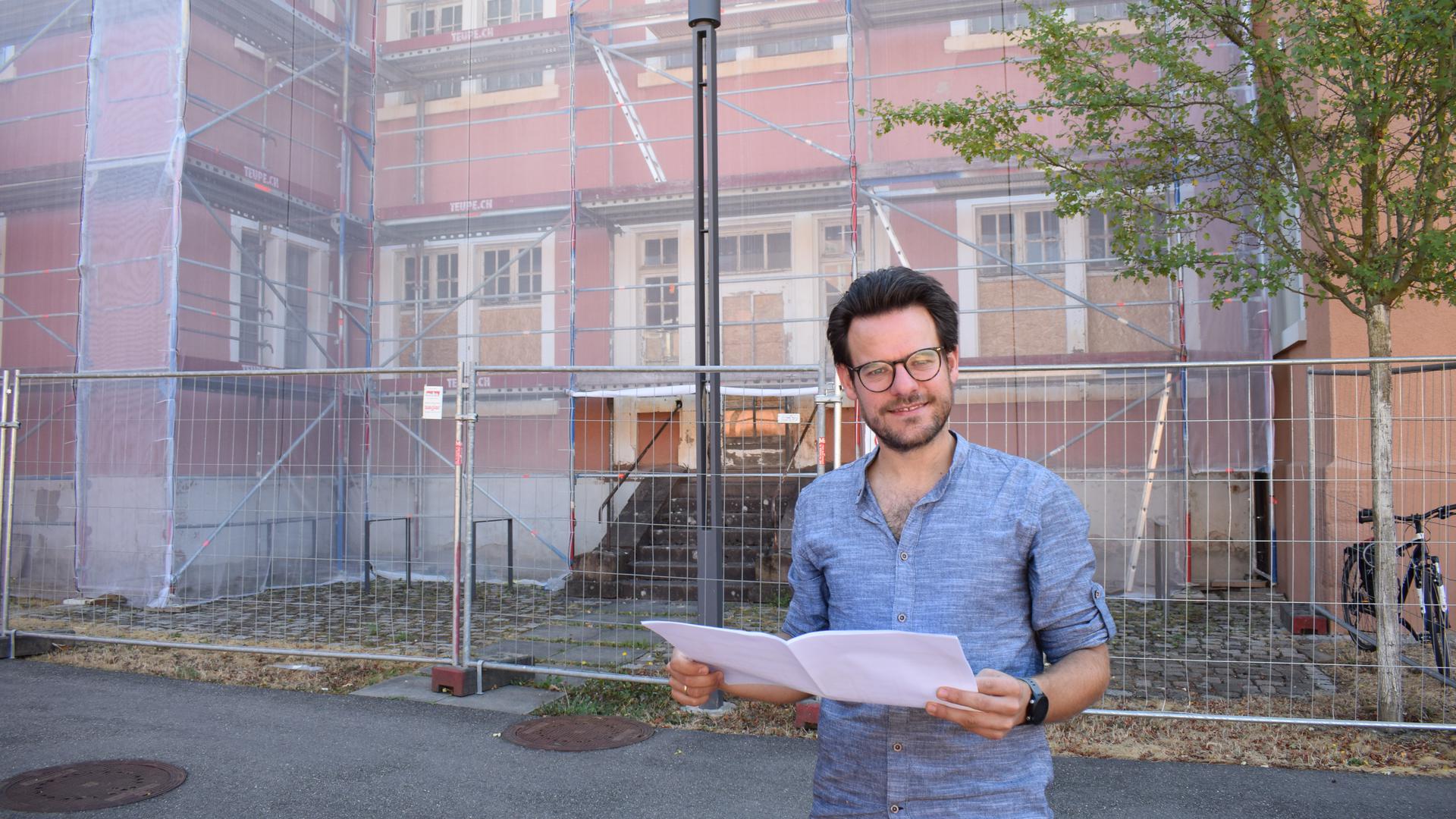 Ein junger Mann mit Brille steht vor einer roten Fassade, die von einem Gerüst umgeben ist.