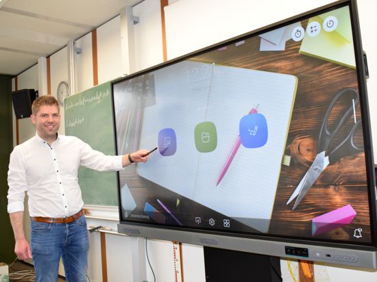 Ein junger Lehrer im weißen Hemd steht vor einem riesigen Bildschirm in einem Klassenzimmer.