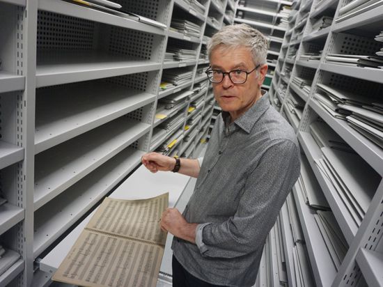 Matthias Kassel steht zwischen Regalen in einem Archiv und blättert in Partituren.