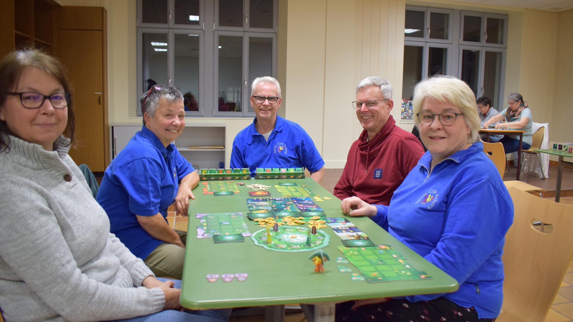 Fünf Erwachsene sitzen um einen grünen Tisch, auf dem ein Spiel aufgebaut ist, und lachen in die Kamera.