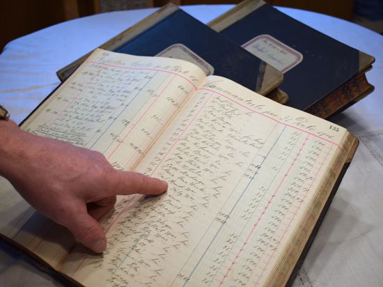 Der Finger eine Männerhand zeigt auf die handgeschriebenen Zeilen eine alten Buches. Dahinter liegen zwei weitere schwarz gebundene Bücher.