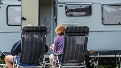 Ein Rentnerehepaar sitzt auf Campingstühlen vor einem Wohnwagen auf einem Campingplatz,