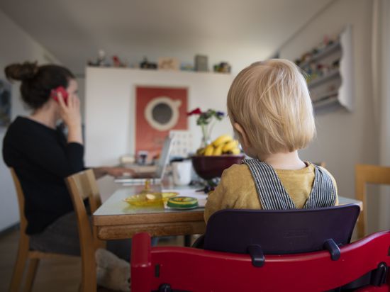 Ein kleines Kind sitzt in einem Hochstuhl vor einem Tisch. Eine Frau sitzt dabei. Sie telefoniert und tippt gleichzeitig auf einem Laptop.