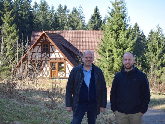 Zwei Männer stehen vor einem Fachwerkgebäude, das von Wald umgeben ist.