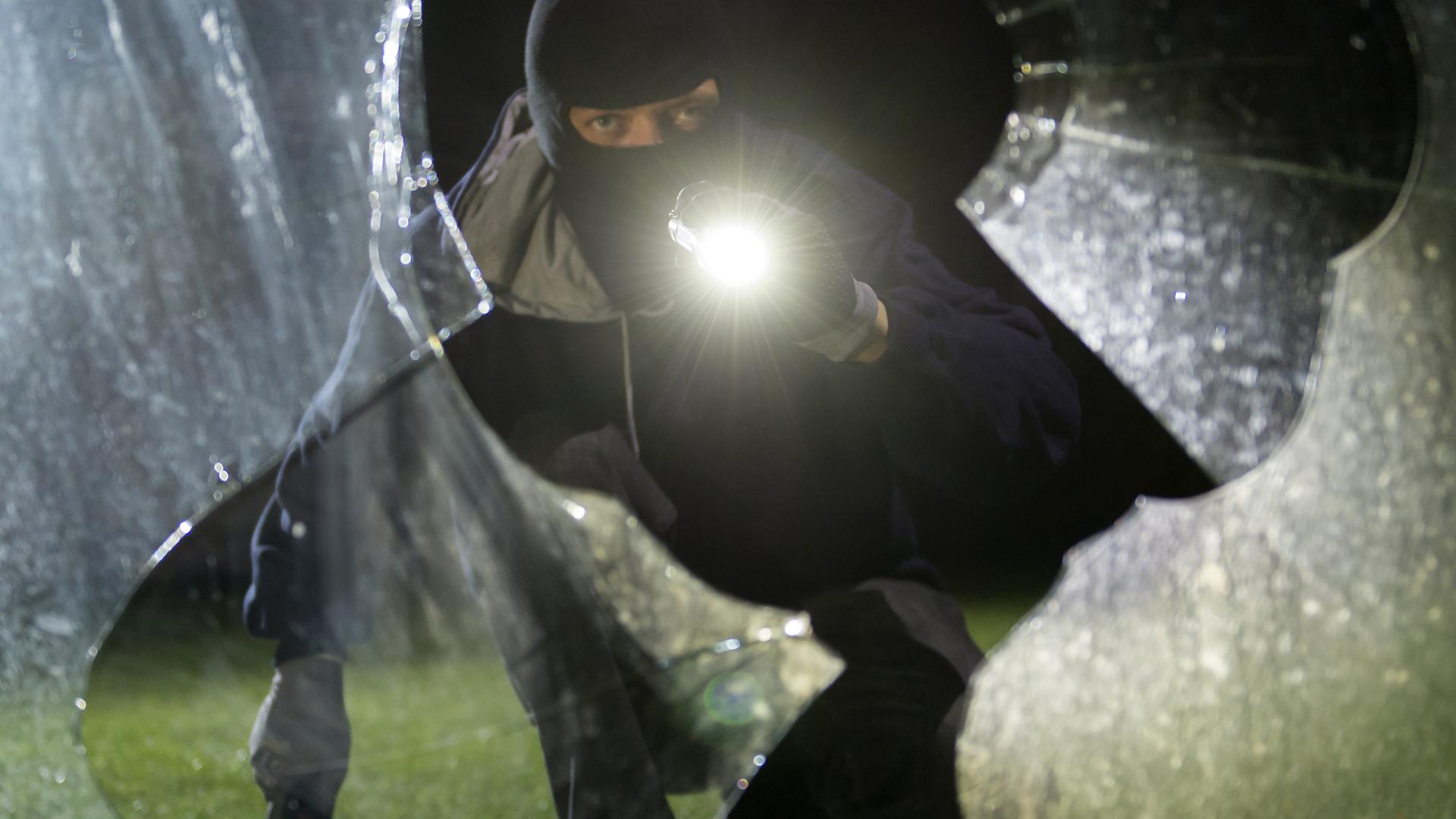 Ein vermummter Einbrecher leuchtet durch eine zerbrochene Scheibe. 
