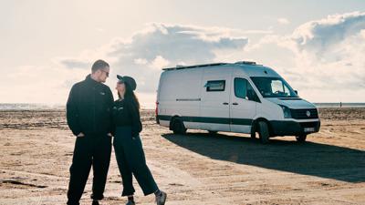 Ein Paar steht bei Sonneuntergang vor seinem umgebauten Van an einem riesigen Sandstrand in Dänemark.