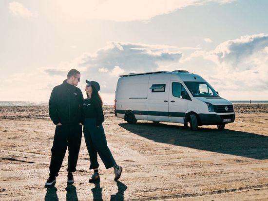 Ein Paar steht bei Sonneuntergang vor seinem umgebauten Van an einem riesigen Sandstrand in Dänemark.