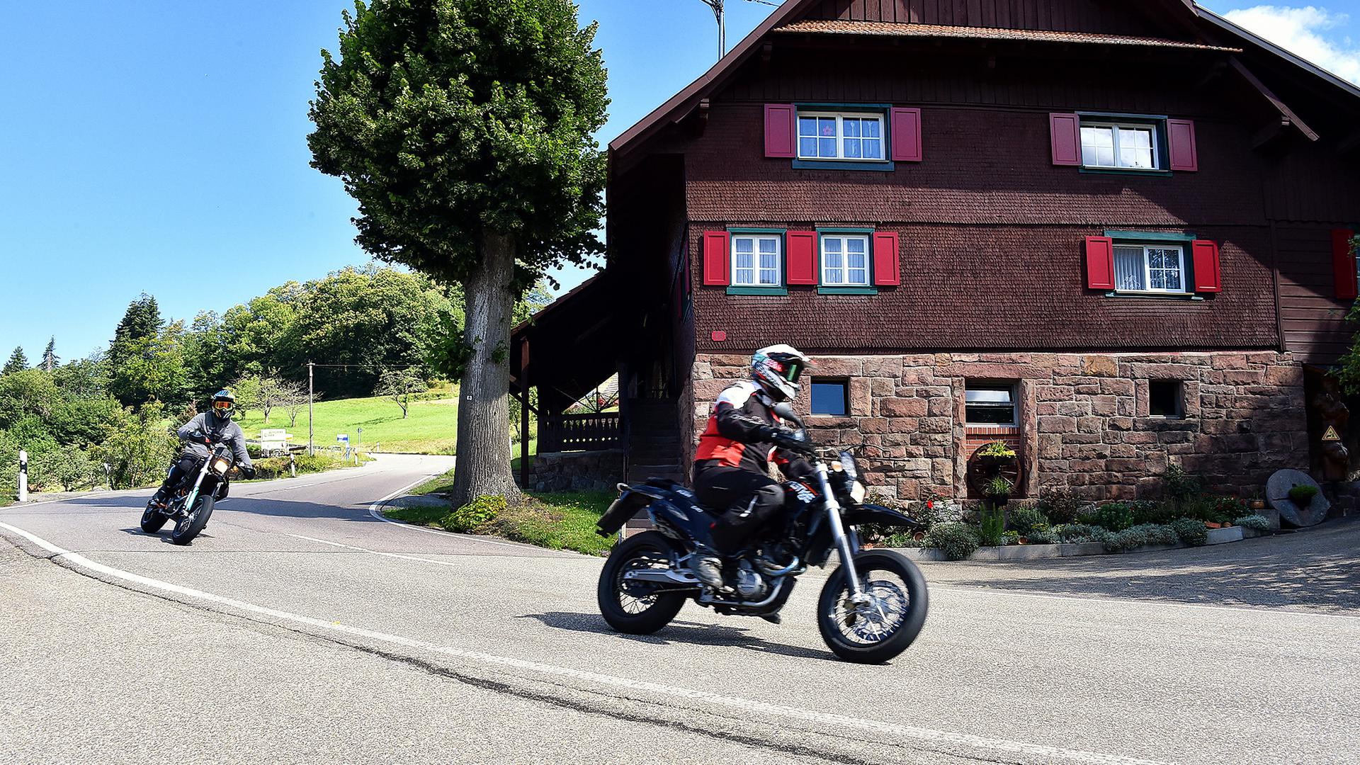 Zwei Motorradfahrer fahren in einer Kurve an einem Haus vorbei.