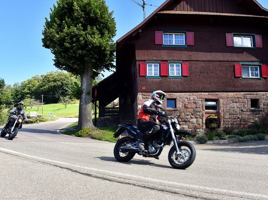 Zwei Motorradfahrer fahren in einer Kurve an einem Haus vorbei.