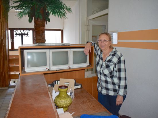 Ein Frau mit Brille steht hinter einem Tresen, an dem alte Bildschirme stehen. Im Hintergrund sind eine künstliche Palme und ein alter Kleiderständer zu sehen.