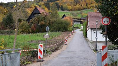 Entlang einer schmalen Straße stehen Warnbacken auf einem mit Schotter befestigten Seitenstreifen. Im Hintergrund steht eine Bauernhaus hinter Bäumen.