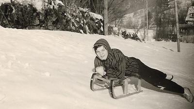 Das Schwarzweißfoto eines Kindes auf einem Schlitten an einem schneebedeckten Hang. 