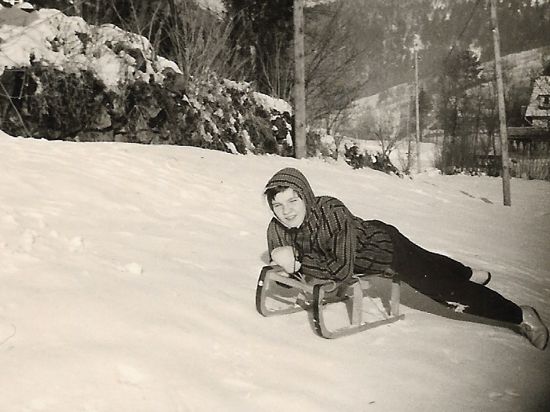 Das Schwarzweißfoto eines Kindes auf einem Schlitten an einem schneebedeckten Hang. 