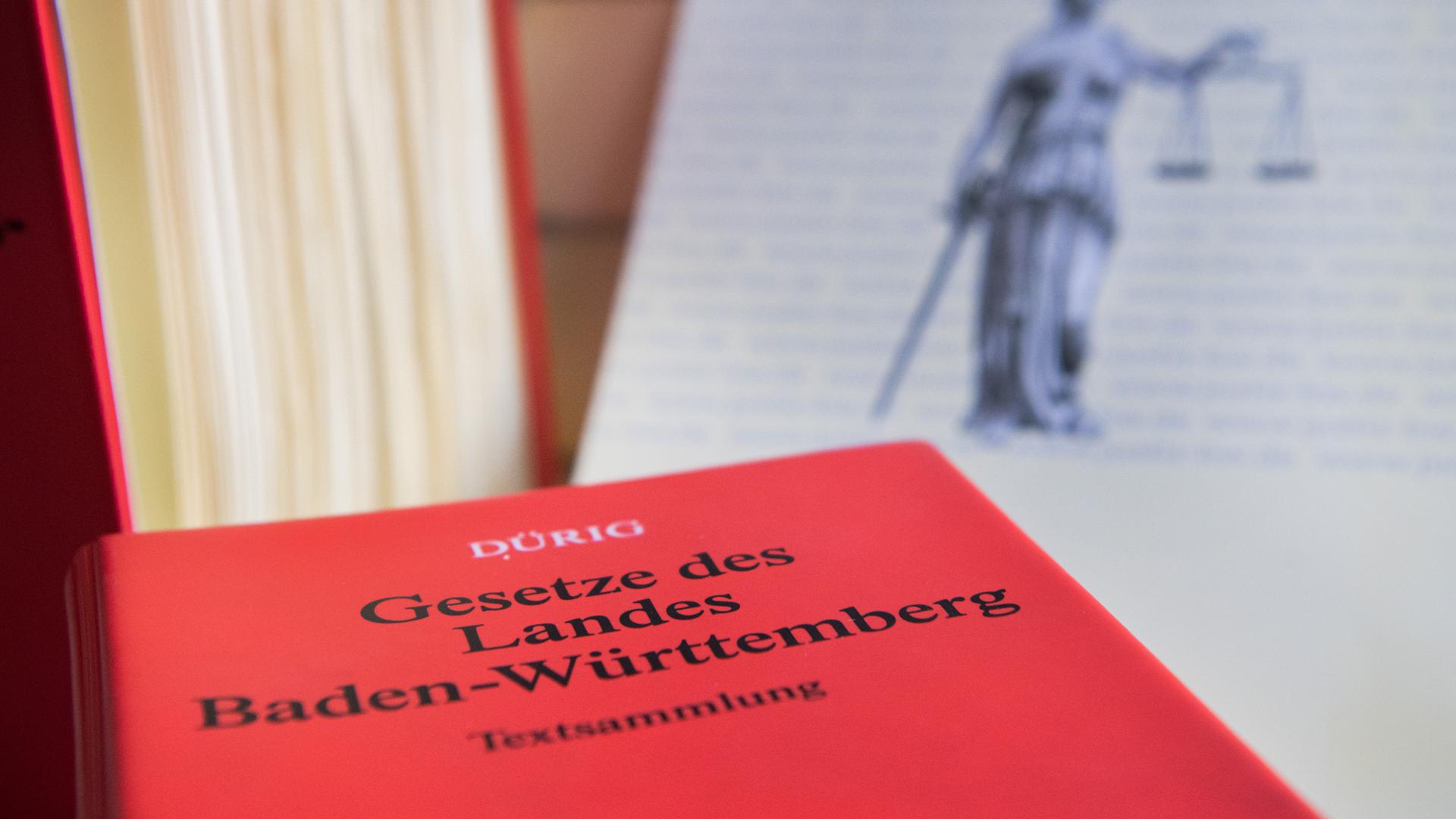 Ein Buch mit der Aufschrift "Gesetze des Landes Baden-Württemberg" liegt auf einem Tisch. Im Hintergrund sind eine Justitia-Abbildung und ein weiteres Gesetzbuch zu sehen (gestellte Szene). 