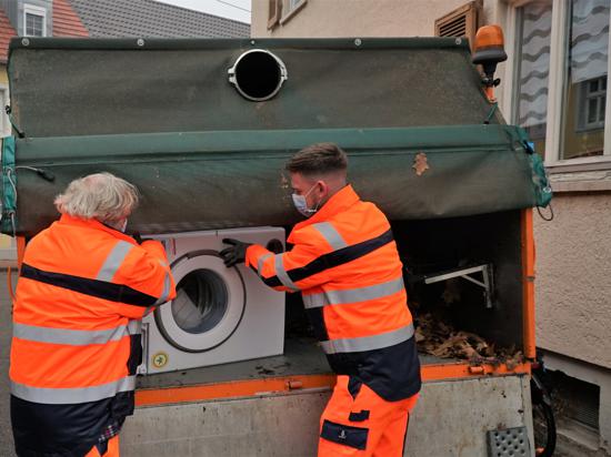 Zwei Männer von der Stadtreinigung laden eine alte Waschmaschine auf einen Lastwagen. 