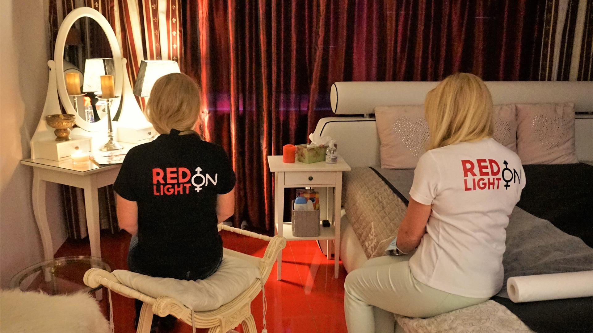 Zwei Damen sitzen mit dem Rücken zum Betrachter auf einem Hocker und auf einem Doppelbett. Sie tragen T-Shirts mit der Aufschrift Redlight on.