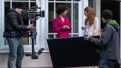 In der Mitte spielen zwei SchauspielerInnen eine Szene, während im Vordergrund ein Crew-Mitglied eine schwarze Platte in den Händen hält und auf der linken Seite ein Kameramann die Szene filmt. 