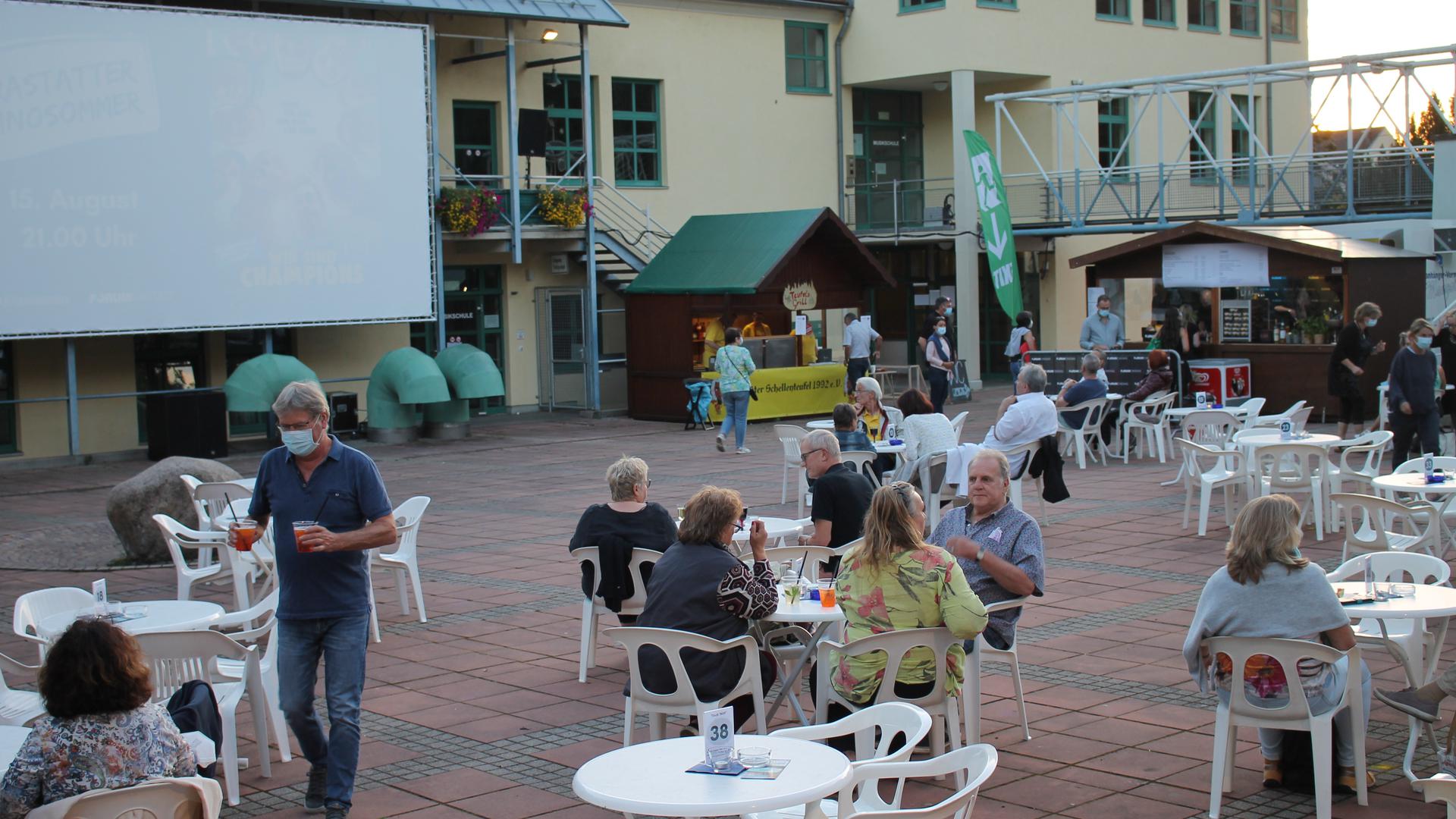 Blick auf eine Freilicht-Kinoleinwand mit Tischen und Menschen davor