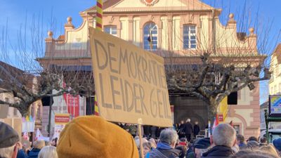 Bei einer Demonstration hält eine Frau ein Schild mit der Aufschrift „Demokratrie - leider geil“ in die Höhe. Vor ihr steht eine Menschenmenge vor dem Rastatter Rathaus. 