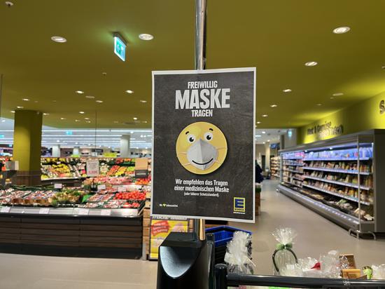 Eingang eines Supermarktes. Schild mit Smiley der einen Mundschutz trägt. Mit den Worten „freiwillig Maske tragen“.