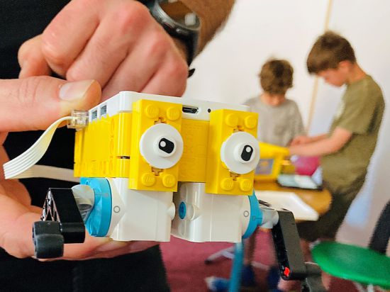 Ein Mann hält einen Lego-Roboter in der Hand. Im Hintergrund programmieren Kinder ein solches Teil. 