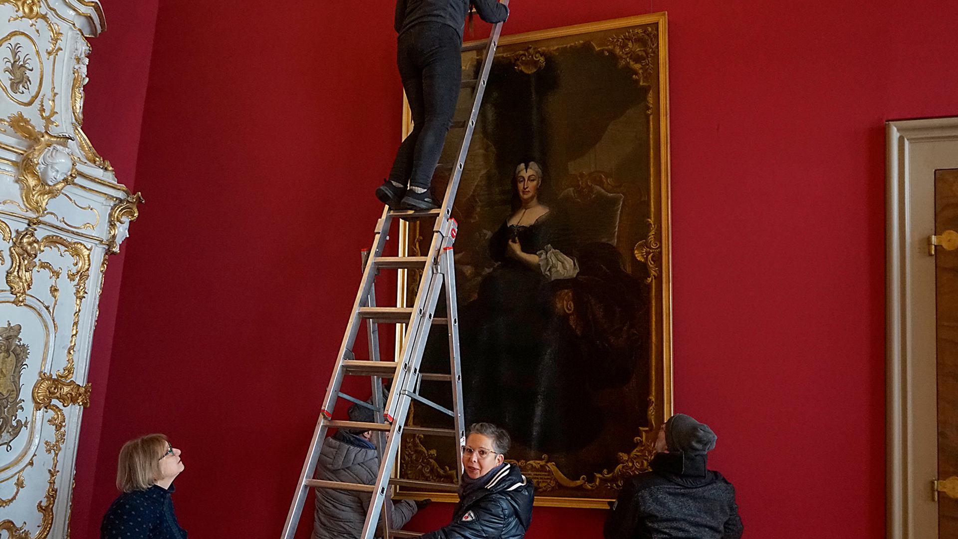 Die Restauratorinnen Juliane Lange (li) und Heidi Kiesslinger (re) und Mitarbeiter der Schlossverwaltung haengen das restaurierte Portrait von Sibylla Augusta an seinen Platz                               