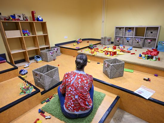 Eine Erzieherin blickt auf einen leeren Gruppenraum einer Kindertagesstätte. Sie ist von hinten zu sehen. Vereinzelt liegt Spielzeug herum.