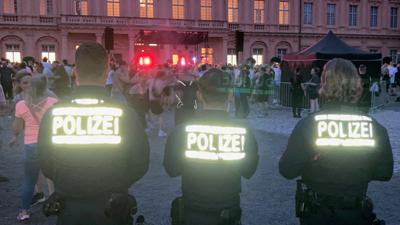 Drei Polizisten stehen mit dem Rücken zur Kamera vor dem Rastatter Schloss beim Elektrofestival Red Residence. Auf ihren Jacken reflektiert die Aufschrift „Polizei“ den Blitz der Kamera.