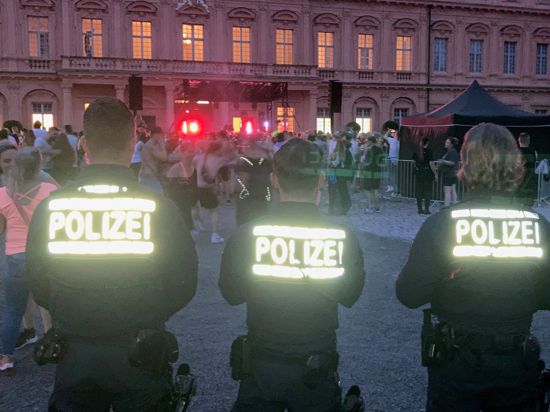 Drei Polizisten stehen mit dem Rücken zur Kamera vor dem Rastatter Schloss beim Elektrofestival Red Residence. Auf ihren Jacken reflektiert die Aufschrift „Polizei“ den Blitz der Kamera.